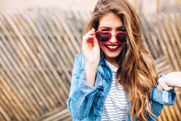 Nahaufnahmeporträt des weißen europäischen lächelnden Mädchens mit langen Haaren und roten Lippen. Attraktive junge lachende Frau ließ stilvolle Sonnenbrille überrascht auf den unscharfen Hintergrund fallen.