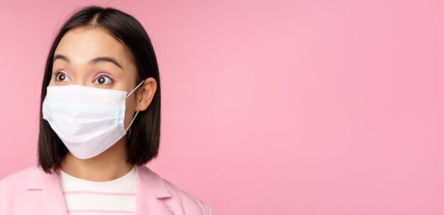 Nahaufnahmeporträt einer asiatischen Geschäftsfrau mit medizinischer Gesichtsmaske, die überrascht aussieht, als sie im Anzug vor rosa Hintergrund steht