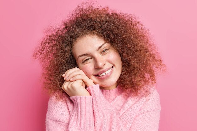 Nahaufnahmeporträt einer erfreuten sanften Frau hält die Hände in der Nähe des Gesichts, lächelt angenehm kippt den Kopf, zeigt zärtlich die Zähne, bewundert etwas, trägt lässige Pullover-Posen drinnen gegen rosa Wand
