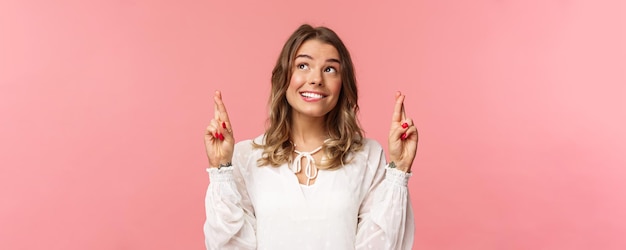 Kostenloses Foto nahaufnahmeporträt einer hoffnungsvollen, optimistischen, attraktiven frau in weißem kleid, die lippe beißt und lächelt, während ein träumender hoffnungstraum wahr wird, der wünsche erfüllt oder einen stehenden rosa hintergrund betet