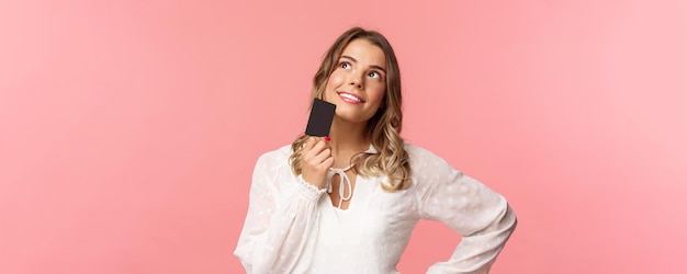 Nahaufnahmeporträt einer nachdenklichen und kreativen blonden Frau in weißem Kleid, die Kinn mit Kreditkarte berührt und verträumt lächelt, während sie nach oben schaut und sich vorstellt, was sie als Geschenk kaufen, rosa Hintergrund