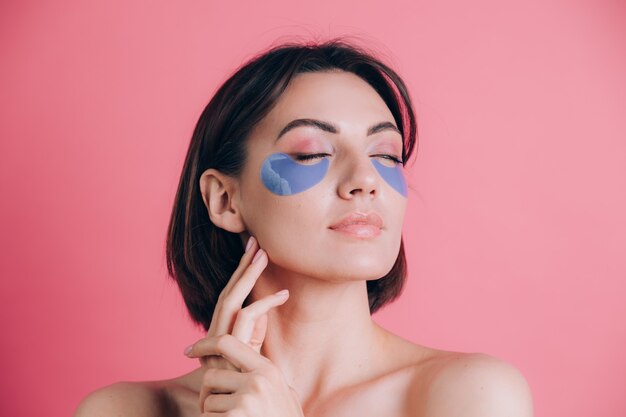 Nahaufnahmeporträt einer schönen jungen Frau oben ohne offene Schultern mit blauen Kollagenpolstern unter ihren Augen. Schönheitskonzept.