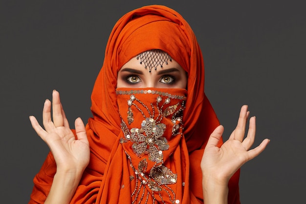 Nahaufnahmeporträt einer süßen jungen Frau mit wunderschönen rauchigen Augen und feinem Schmuck auf der Stirn, die den mit Pailletten verzierten Terrakotta-Hijab trägt. Sie sieht erschrocken auf einem dunklen Hintergrund aus. Summen