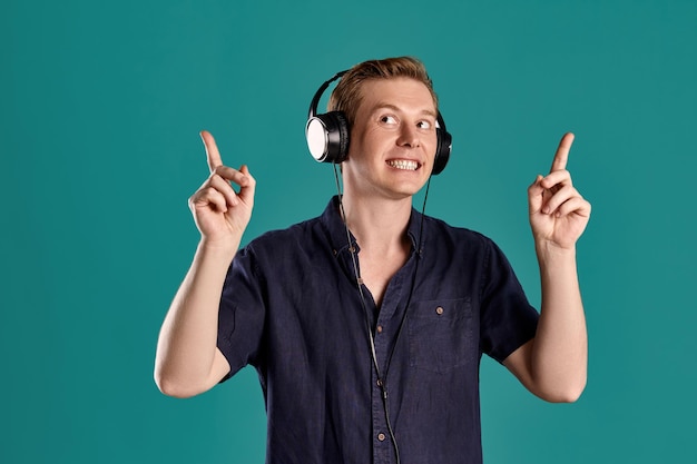 Nahaufnahmeporträt eines erwachsenen gutaussehenden rothaarigen Kerls in einem stylischen marineblauen T-Shirt, der die Musik über Kopfhörer hört, während er auf blauem Studiohintergrund posiert. Menschliche Gesichtsausdrücke. Aufrichtige Emotionen