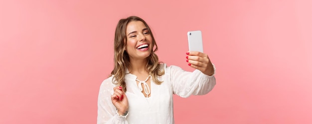 Nahaufnahmeporträt eines fröhlichen, optimistischen, lächelnden blonden Mädchens, das ein weißes Kleid trägt und lacht, als ein Rekord-Videoanruffreund auf einer mobilen Anwendung aufnimmt, die ein Foto-Selfie auf dem rosa Hintergrund des Smartphones macht
