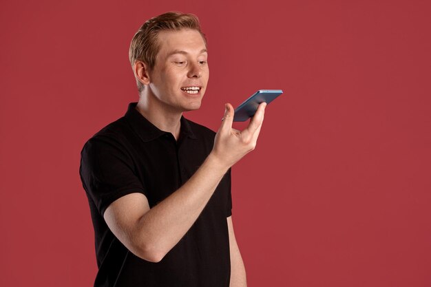 Nahaufnahmeporträt eines jungen, eleganten rothaarigen Mannes in einem stilvollen schwarzen T-Shirt, der mit einem Smartphone spricht, während er auf rosafarbenem Studiohintergrund posiert. Menschliche Gesichtsausdrücke. Aufrichtiges Gefühlskonzept. Leerzeichen kopieren