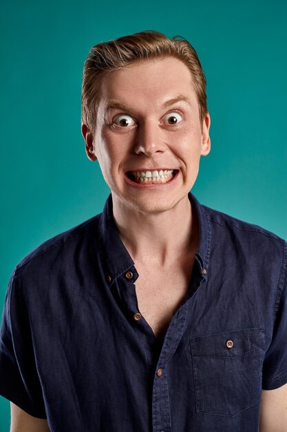 Nahaufnahmeporträt eines jungen netten rothaarigen Kerls in einem stylischen marineblauen T-Shirt, der wütend aussieht, während er auf blauem Studiohintergrund posiert. Menschliche Gesichtsausdrücke. Aufrichtiges Gefühlskonzept. Platz kopieren.