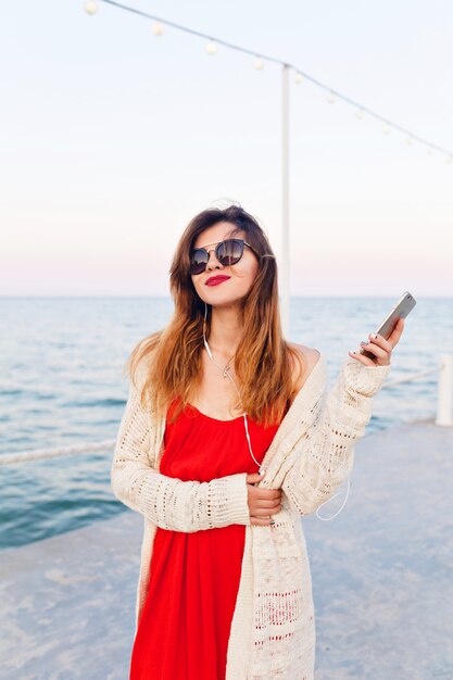 Nahaufnahmeporträt eines schönen Mädchens im roten Kleid und in der weißen Jacke auf einem Pier, lächelnd und Musik auf Kopfhörern auf einem Smartphon hörend