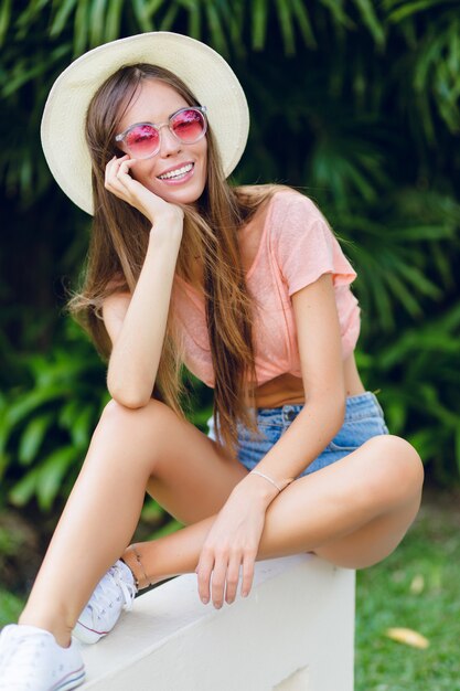 Nahaufnahmeporträt eines schönen stilvollen lächelnden Mädchens, das auf weißem Zaun im tropischen Park sitzt