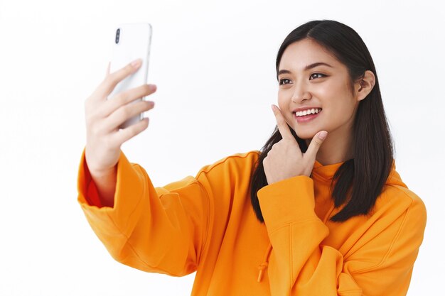 Nahaufnahmeporträt eines stilvollen modernen asiatischen Mädchens in orangefarbenem Hoodie, das Selfie mit dem Handy macht, als Videoaufnahme posiert und lächelt, Blogger, der neue Fotofilter anprobiert, weiße Wand stehend