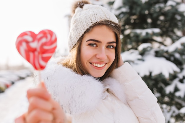 Nahaufnahmeporträt erstaunliche freudige lächelnde Frau im sonnigen Wintermorgen mit rosa lollypop auf Straße. Attraktive junge Frau im weißen warmen Wollhut, der kaltes Wetter genießt. Glückliche Zeit, positive Emotionen.