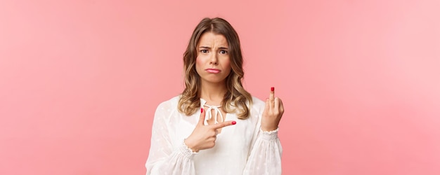 Nahaufnahmeporträt trauriges und frustriertes junges blondes Mädchen wartete auf Vorschlag während eines romantischen Dates, das auf den Finger ohne Hochzeitsring mit verwirrtem verärgertem Ausdruck auf rosa Hintergrund zeigt