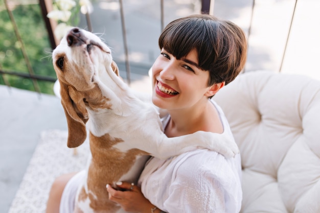 Nahaufnahmeporträt von oben des lachenden Mädchens, das Morgen auf Balkon mit lustigem Haustier genießt. Schöne junge Dame in guter Laune, die mit Beagle-Hund spielt, während sie sich nach dem Abendessen auf der Terrasse ausruht