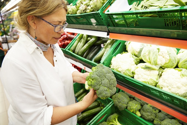 Nahaufnahmeprofilporträt des gutaussehenden Vegetariers der kaukasischen Frau mittleren Alters in der Freizeitkleidung, die das frischeste Gemüse und den frischesten Brokkoli im Lebensmittelgeschäft aufnimmt und auswählt. Menschen und Einkaufen