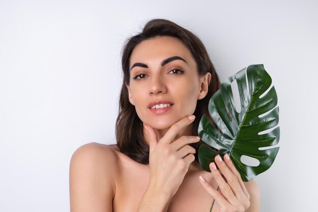 Nahaufnahmeschönheitsporträt einer schulterfreien Frau mit perfekter Haut und natürlichem Make-up mit Monstera-Palmblatt