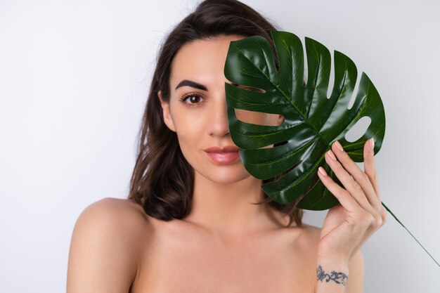 Nahaufnahmeschönheitsporträt einer schulterfreien Frau mit perfekter Haut und natürlichem Make-up mit Monstera-Palmblatt