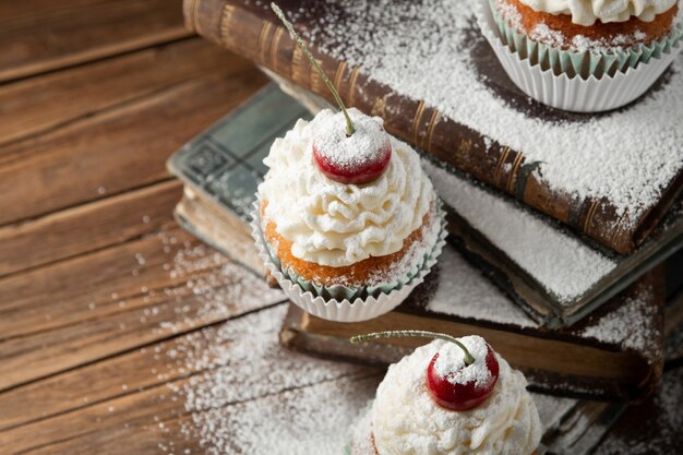 Nahaufnahmeschuss von köstlichen Cupcakes mit Sahne, Puderzucker und einer Kirsche oben auf Büchern