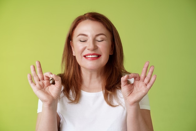 Kostenloses Foto nahe an der perfektion nahaufnahme friedlich entspannt rothaarige glückliche frau geschlossene augen reines entzücktes lächeln zeigen zen frieden zufriedenheit geste meditation reichweite nirvana ruhe stehen grüne wand