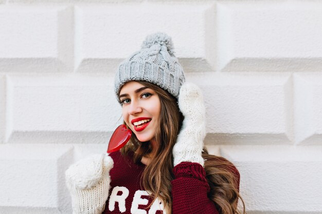 Nahes hübsches Mädchen mit langen Haaren im Marsala-Pullover auf grauer Wand. Sie trägt weiße Handschuhe, eine graue Strickmütze, hält einen roten Herzlutscher und lächelt.