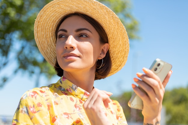 Nahes Porträt der Frau im Freien im gelben Sommerkleid und im Hut am sonnigen Tag halten Handy und schauen nach links