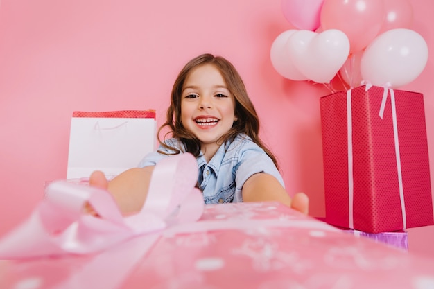 Nahes rosa Geschenk, das kleines freudiges Mädchen zur Kamera auf rosa Hintergrund gibt. Lächelnde große Geschenkboxen, Luftballons, Geburtstagsfeier feiern, Positivität ausdrücken