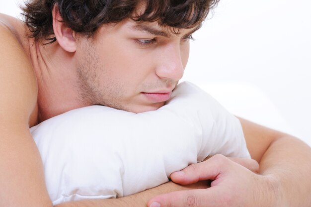Nahes trauriges Gesicht eines jungen kaukasischen Mannes, der im Bett liegt und das Kissen umarmt