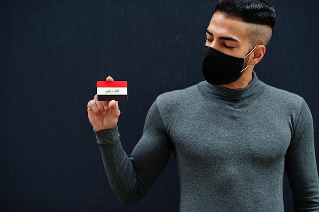 Nahöstlicher Mann in grauem Rollkragenpullover und schwarzer Gesichtsschutzmaske zeigt isolierten Hintergrund der irakischen Flagge