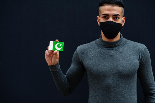 Nahöstlicher Mann in grauem Rollkragenpullover und schwarzer Gesichtsschutzmaske zeigt pakistanische Flagge isolierten Hintergrund