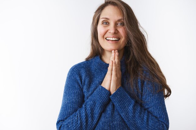 Namaste-Frau mittleren Alters, die sich entspannt und glücklich fühlt und die Handflächen in der Nähe des Kinns zusammengepresst hält, betet, lächelt freundlich und dankt für die Hilfe, die dankbar ist und in einem blauen Pullover auf weißem Hintergrund posiert