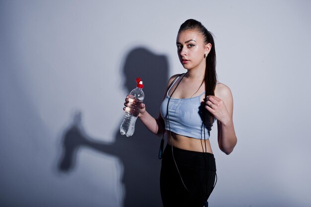 Nasse, fröhliche, attraktive junge Fitness-Frau in Top und schwarzen Leggings mit Springseil und Flasche Wasser isoliert auf weißem Hintergrund