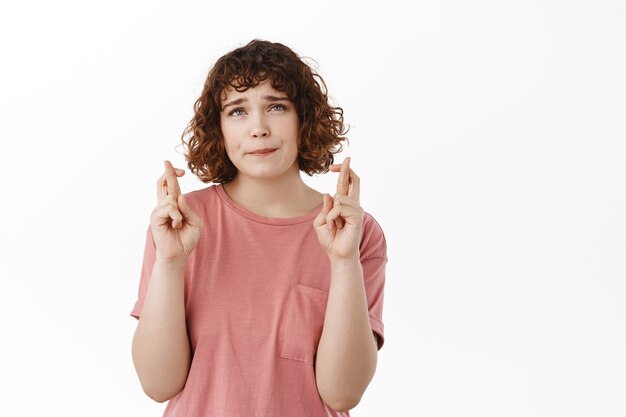 Nervöse besorgte Studentin, Daumen drücken und aufblicken, um Wünsche zu machen, für einen Traum zu beten, auf ein Wunder zu warten, auf Weiß zu stehen