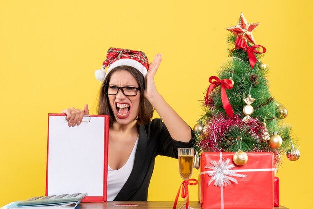 Nervöse charmante Dame im Anzug mit Weihnachtsmannhut und Brille, die Dokument im Büro zeigt