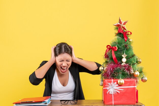 Nervöse junge Frau im Anzug nahe geschmücktem Weihnachtsbaum im Büro