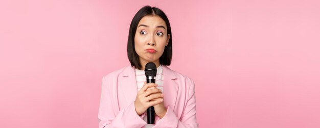 Nervöse Unternehmerin, die eine Rede mit Mikrofon hält und ängstlich zur Seite schaut, posiert im Anzug vor rosa Hintergrund