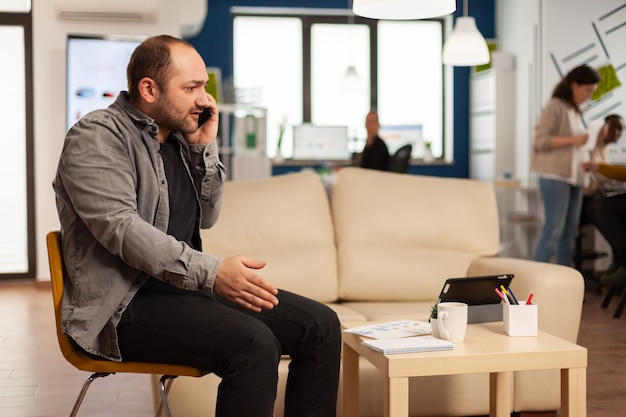 Nervöser Manager, der aggressiv telefoniert und flucht, schreiend auf einem Stuhl mitten im Geschäftsbüro sitzt