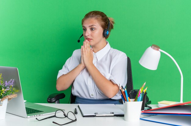 Nervöses junges blondes Call-Center-Mädchen mit Headset am Schreibtisch sitzend mit Arbeitswerkzeugen, die die Hände zusammenhalten und auf den Laptop schauen, der auf grüner Wand isoliert ist