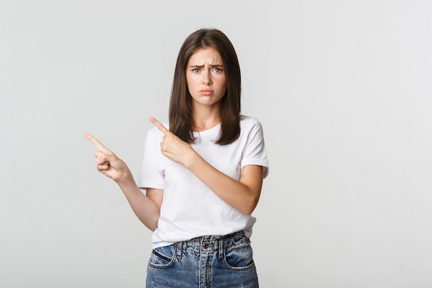 Nervöses schüchternes süßes Mädchen, das Finger oben links zeigt und traurig aussieht, Bedauern habend.