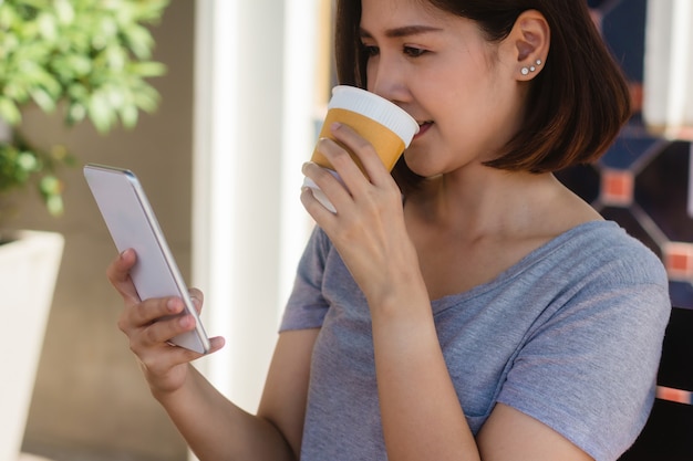 Nette asiatische junge Frau, die in trinkendem Kaffee des Cafés sitzt und Smartphone für die Unterhaltung verwendet