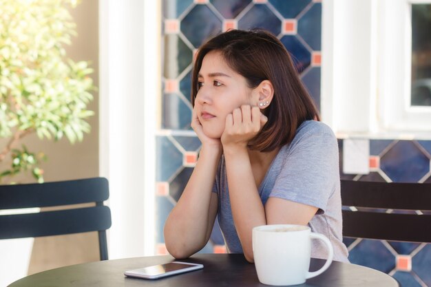 Nette asiatische junge Frau, die in trinkendem Kaffee des Cafés sitzt und Smartphone für die Unterhaltung verwendet