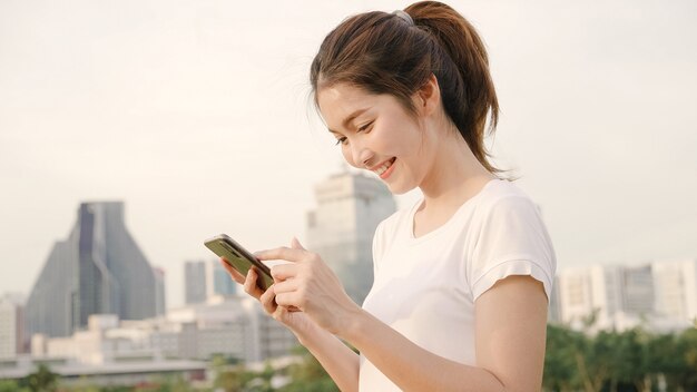 Nette asiatische touristische Bloggerfrau, die Smartphone für Richtung verwendet und auf Standortkarte beim Reisen auf die Straße in die im Stadtzentrum gelegene Stadt schaut.