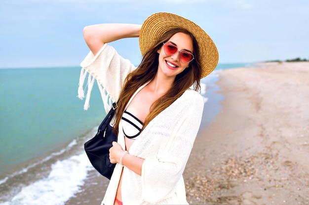 Nette blonde Frau, die Selfie am Ozeanstrand macht, Boho-Outfit und lustige Sonnenbrille tragend, Vintage Strohhut, Kuss senden.