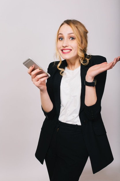 Nette blonde junge Bürofrau im weißen Hemd, schwarzer Anzug mit Telefon, das lokal schaut. Ausdruck wahrer positiver Emotionen, Erfolg, Arbeit, Freundlichkeit