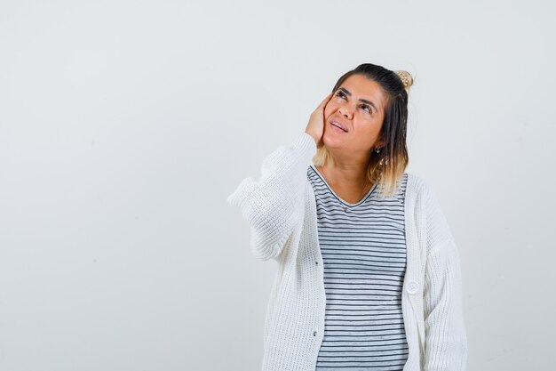 Nette Dame, die Hand auf Wange hält, in T-Shirt, Strickjacke nach oben schaut und hoffnungsvoll aussieht, Vorderansicht.