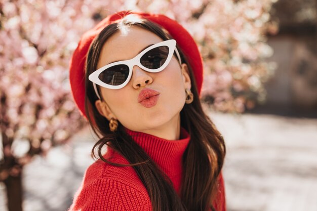 Nette Dame in Baskenmütze und Sonnenbrille bläst Kuss gegen Hintergrund von Sakura. Attraktive stilvolle Frau im roten Pullover, der kokett im Garten aufwirft