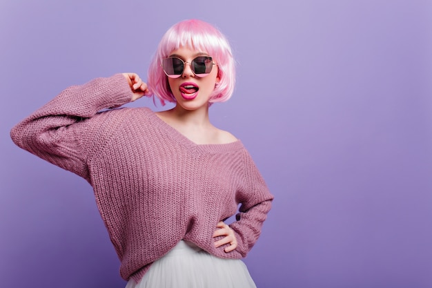 Nette europäische Frau in der bunten Perücke, die mit Zunge heraus aufwirft. selbstbewusstes glückseliges Mädchen mit rosa Haaren, die Brille und lila Pullover tragen.