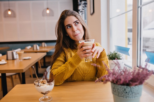 Nette Frau im trendigen gelben Pullover, der an etwas denkt, während im Café mit Glas Cappuccino ruht. Innenporträt der atemberaubenden Dame, die auf Freund wartet und Eis genießt.