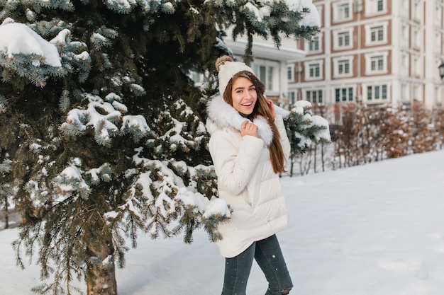 Nette Frau im trendigen weißen Mantel, der Spaß während des Winterfotoshootings und des Lachens hat. Außenfoto der prächtigen brünetten Dame trägt lustigen Hut im kalten sonnigen Tag.