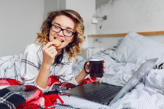 Nette Frau mit gewellter Frisur, die Kekse mit Kaffee isst, Video auf ihrem Laptop schauend.