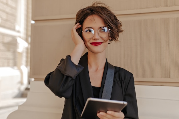 Nette Frau mit kurzen Haaren und roten Lippen, die draußen lächeln. Fröhliche Frau mit rotem Lippenstift in schwarzer Kleidung und Brille hält Tablette im Freien.