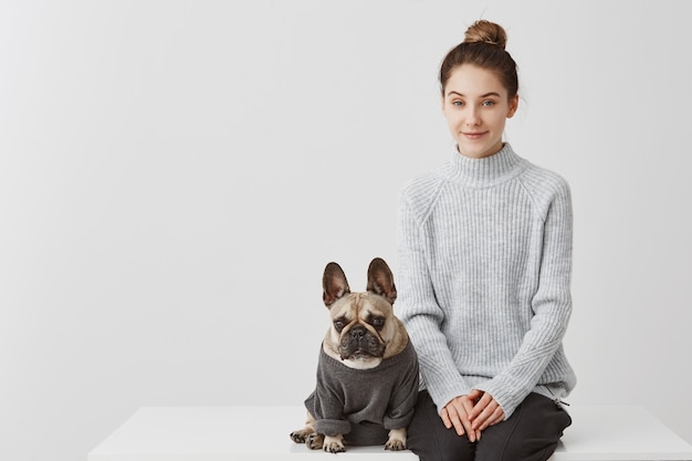 Kostenloses Foto nette frau mit modefrisur und ihrem welpen französische bulldogge gekleidet im pullover. weibliches modell, das auf tisch mit hund über weißer wand sitzt. freundschaftskonzept, kopierraum